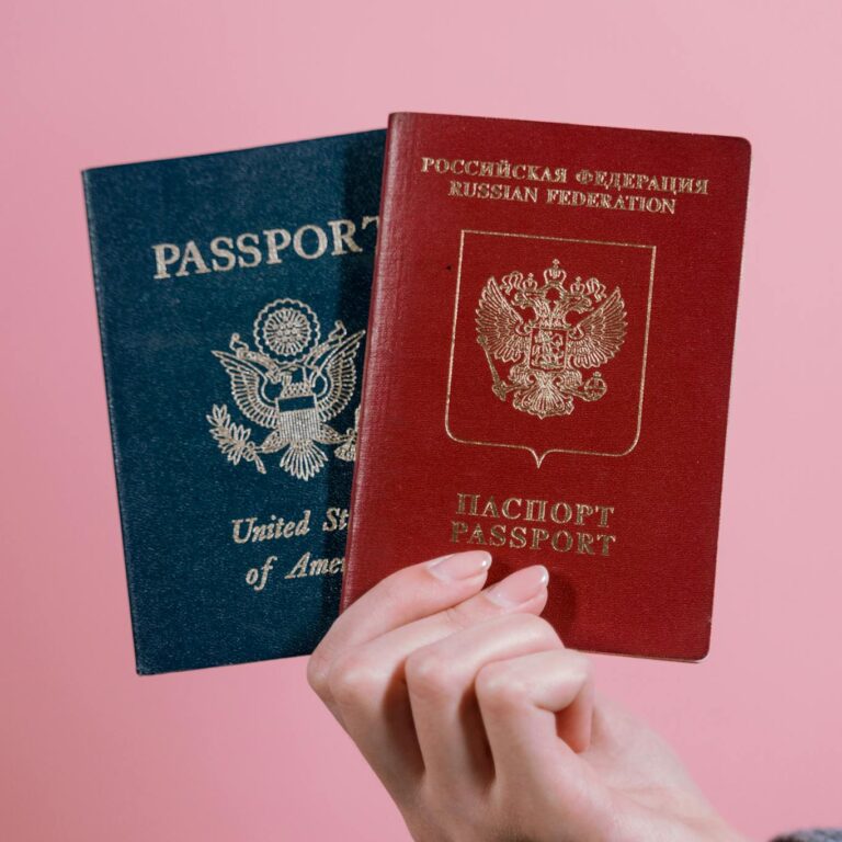 A hand holding US and Russian passports against a pink background.