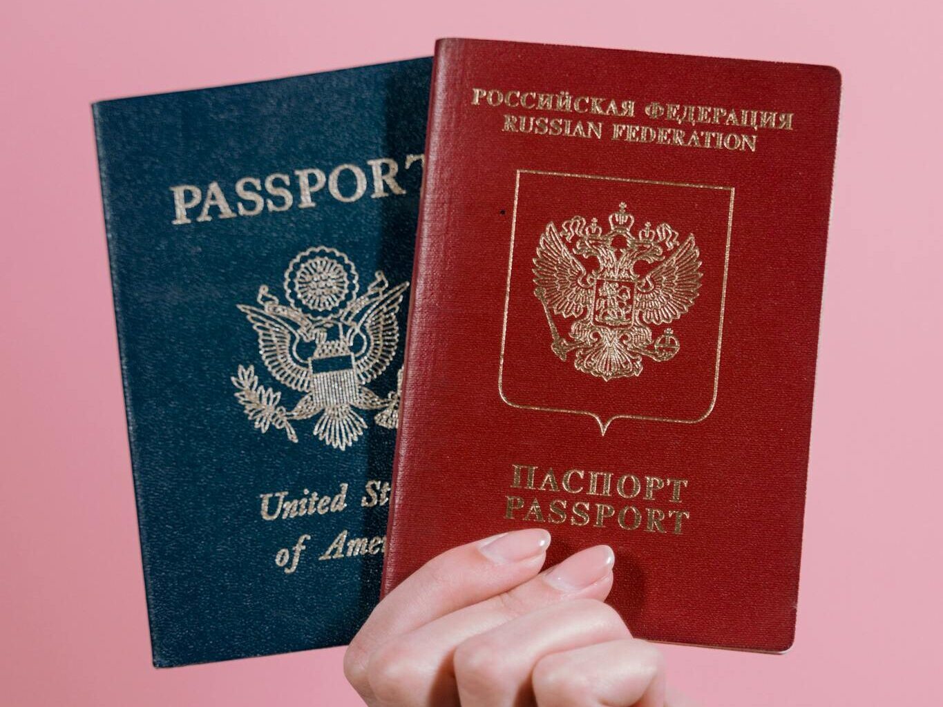 A hand holding US and Russian passports against a pink background.